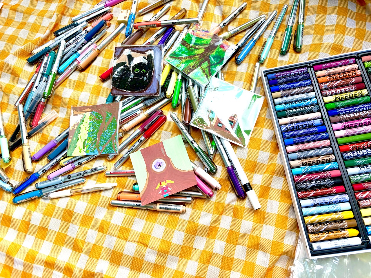 Acrylic markers and oil pastels surrounding small artworks on a yellow checkered blanket